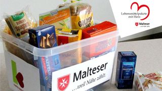 Transparente Kunststoffbox, gefüllt mit Lebensmitteln, darauf ein Aufkleber „Malteser ...weil Nähe zählt“, daneben ein Päckchen Kaffee