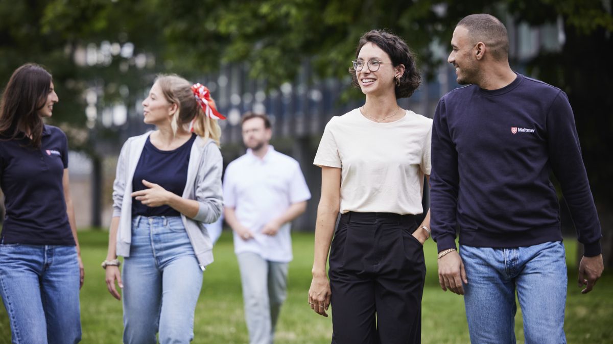 Eine Gruppe junger Malteser Mitarbeitenden, jeglichen Aussehens und Geschlechts, schlendern lachend über eine Grünfläche.