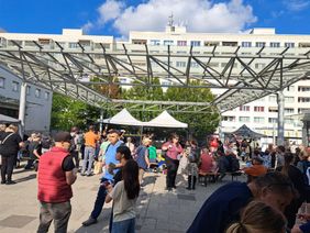 Viele Menschen genießen das Bürgerfest unter freiem Himmel. Im Hintergrund ist ein Wohnblock von Prohlis zu sehen. Die Sonne scheint.