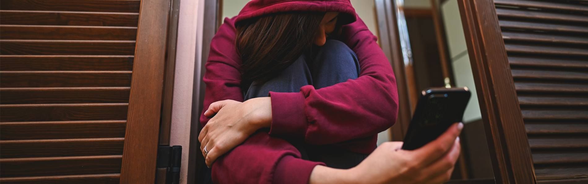 Eine junge Frau mit einer Kapuze über dem Kopf sietzt und schaut auf ein Smartphone 