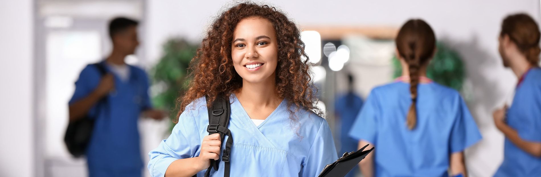 Frau in blauer Kasackkleidung mit Rucksack und Klemmbrett im Klinikflur.