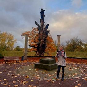 Eine Frau steht in einem Park. Sie liest etwas von einem Blatt.