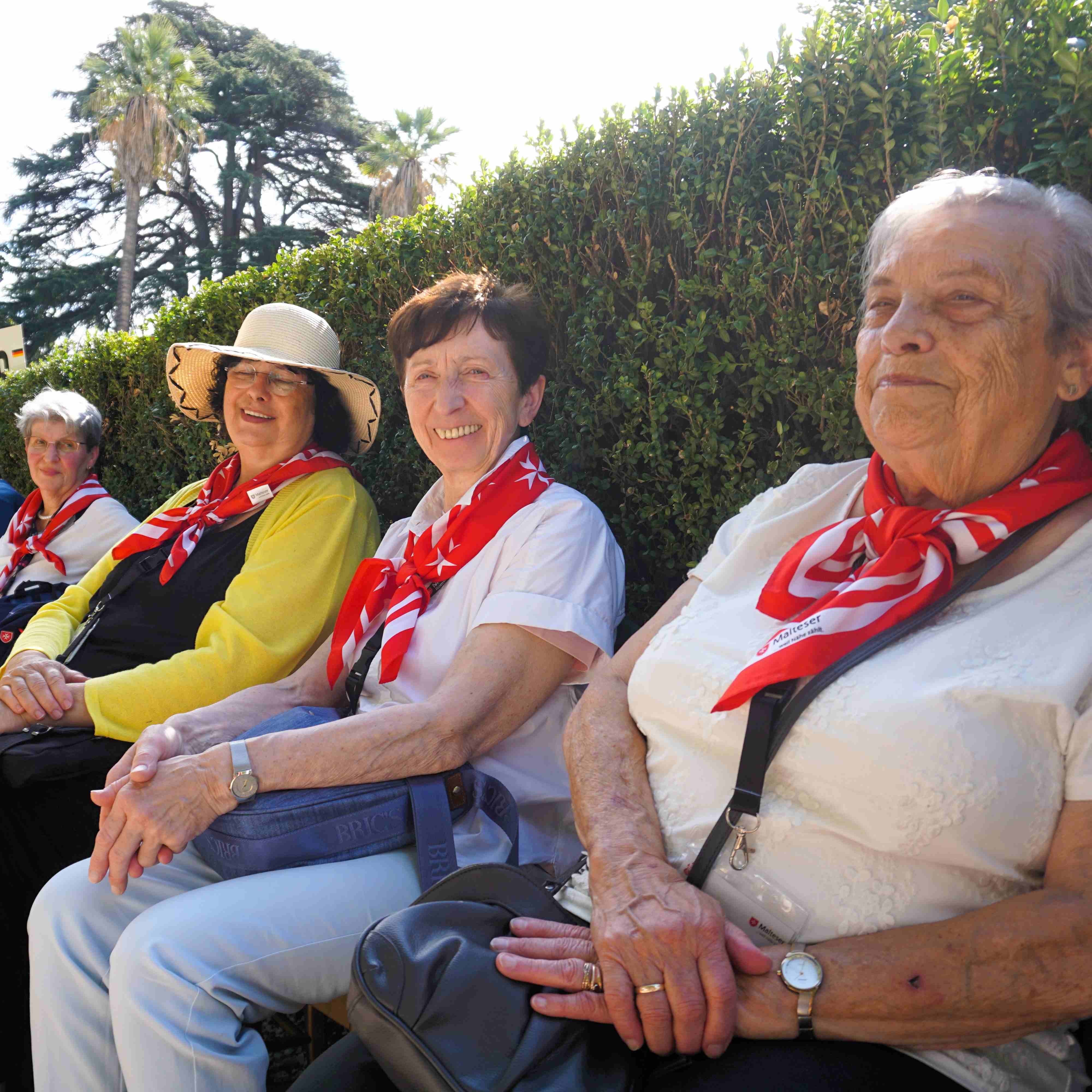 Vier sitzende Personen mit roten Halstüchern, eine trägt einen gelben Pullover, im Hintergrund Hecke und Bäume.
