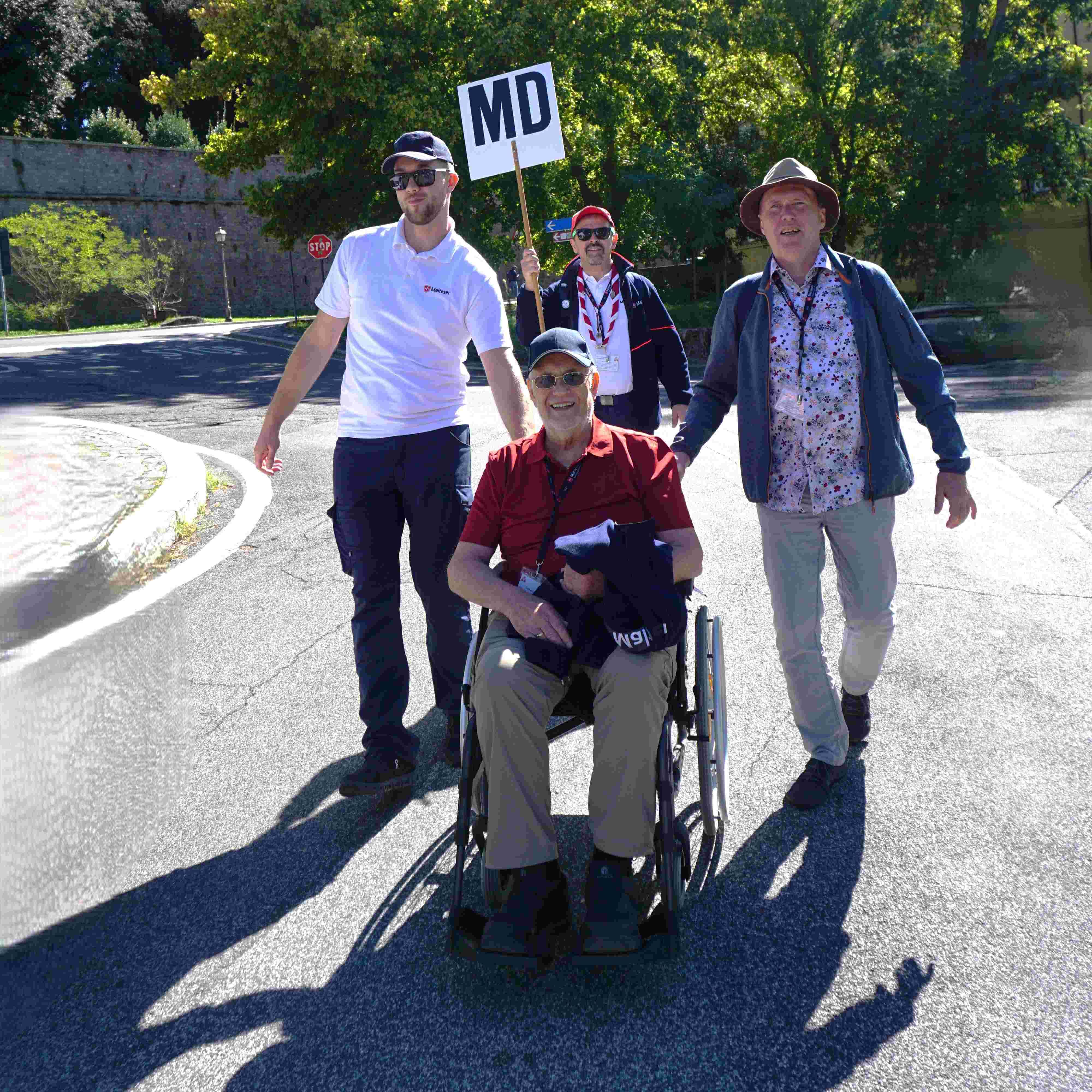 Person im Rollstuhl wird von drei Personen auf einer Straße begleitet, eine Person hält ein Schild mit der Aufschrift 'MD'.