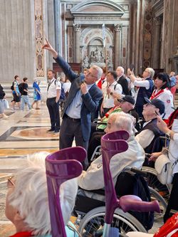Eine Menschenmenge sieht nach oben, während ein Touristguide nach oben zeigt. Einige der Menschen sitzen im Rollstuhl oder haben Gehhilfen.