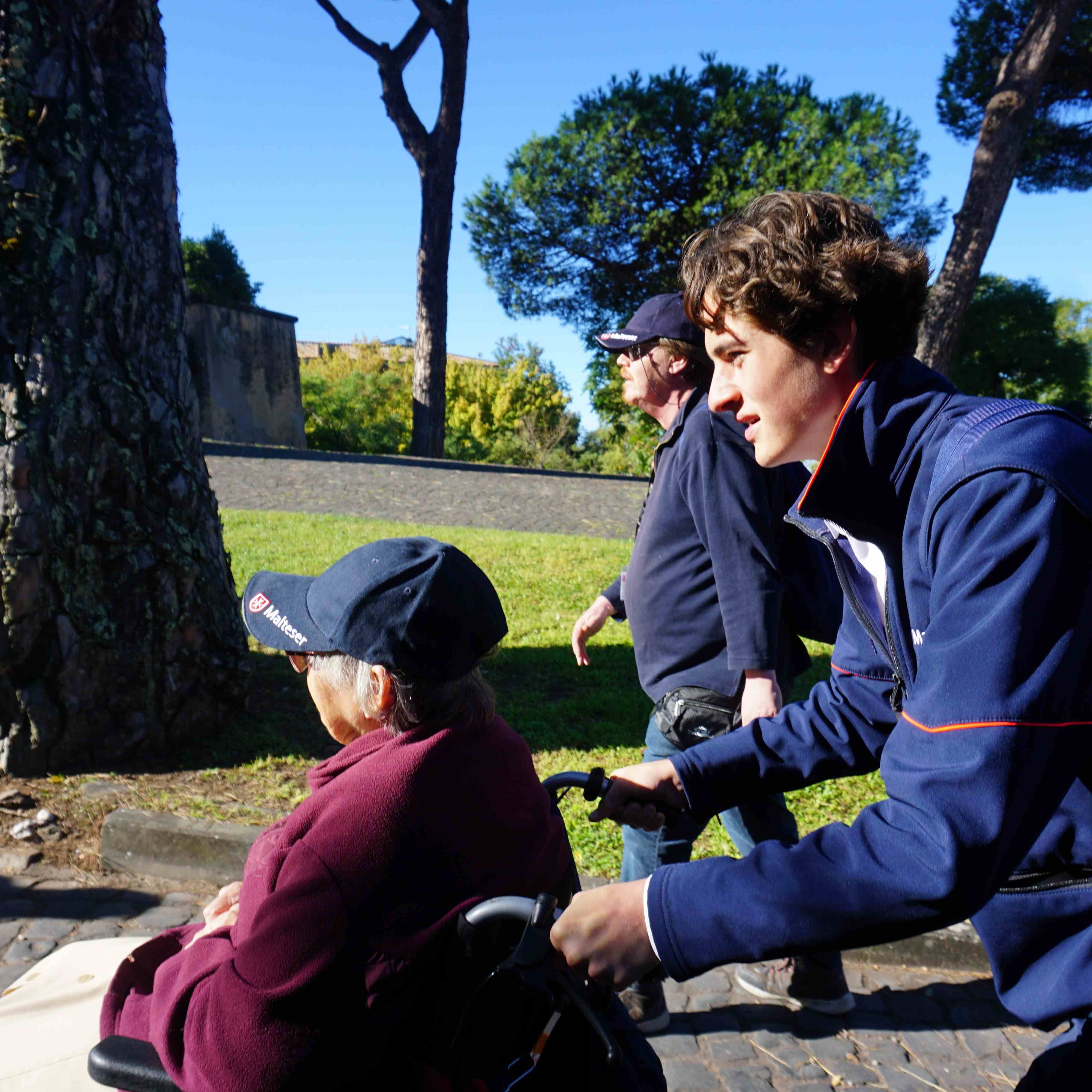 Person im Rollstuhl mit dunkelblauer Mütze und rotem Pullover, begleitet von zwei Personen in blauer Kleidung, im Park mit Bäumen und Wiese.