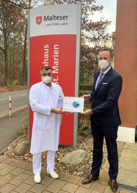 Arzt und Mann im Anzug stehen vor einem Malteser Schild und halten gemeinsam eine Urkunde.