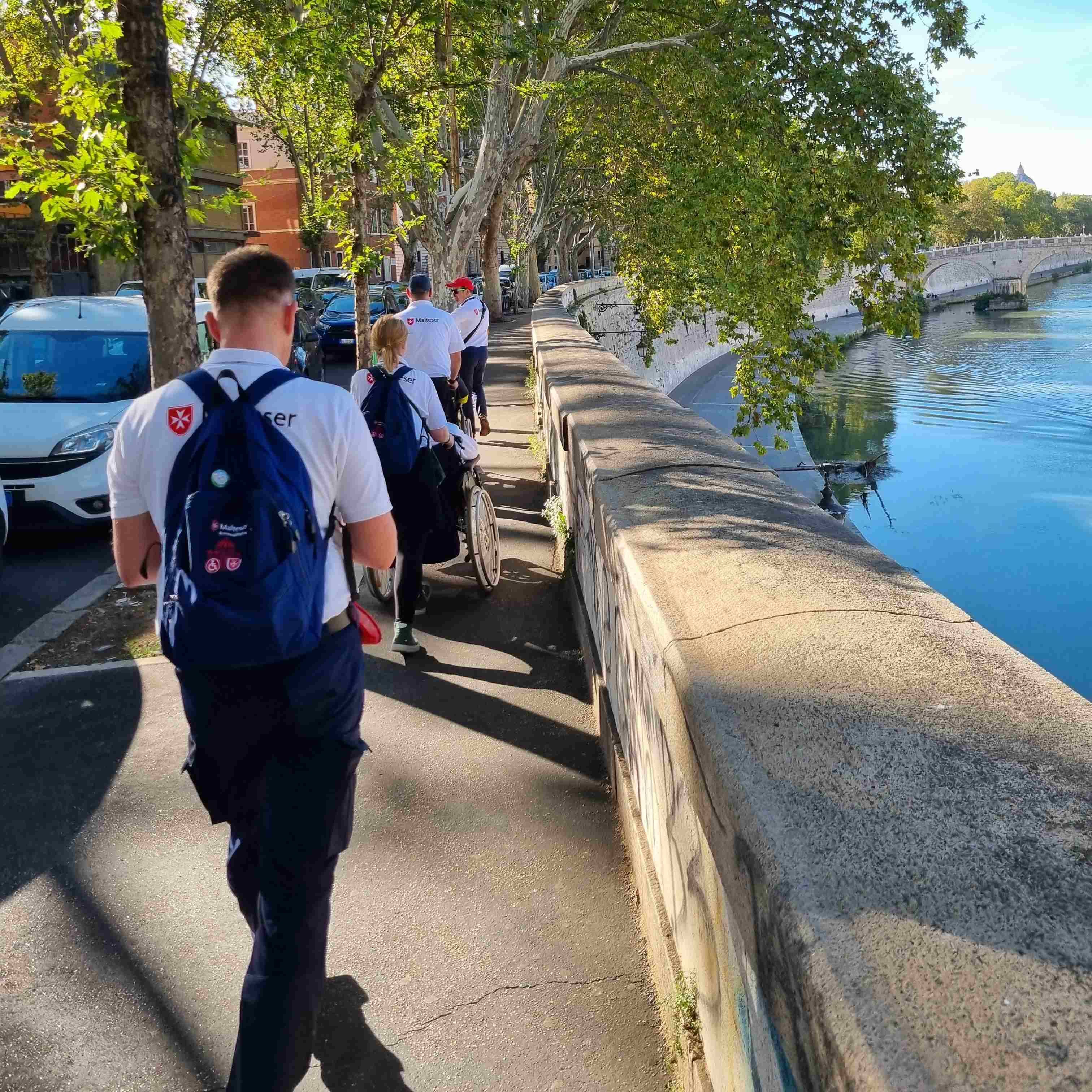 Personen in Malteser Kleidung mit Rucksäcken gehen an einer Ufermauer entlang eines Flusses bei Sonnenschein.