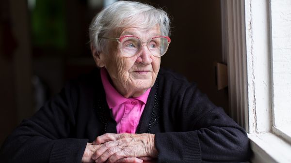 Eine alte Frau mit Brille schaut aus einem dunklen Raum aus einem Fenster und lächelt leicht.