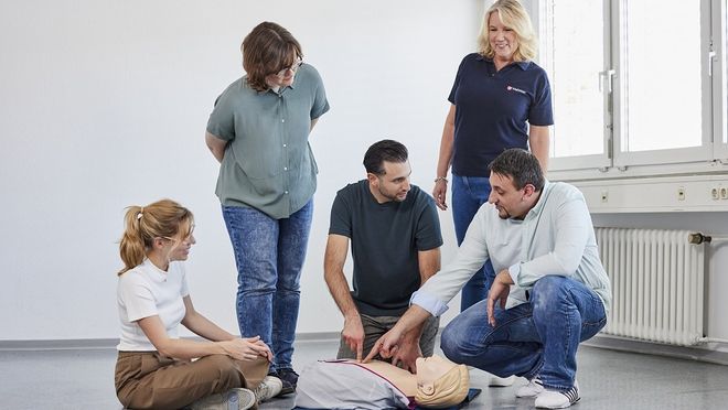 Ein Mann kniet neben einer Ausbildungspuppe und zeigt auf einen Punkt des Oberkörpers, eine Malteser Ausbilderin und weitere Personen sitzen und stehen um ihn herum.