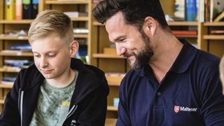 Mann mit dunklem Bart und blauem Malteser Polohemd sitzt lächelnd neben einem blonden Jungen, der etwas schreibt. Hinter den beiden ein Regal mit Lernmaterialien