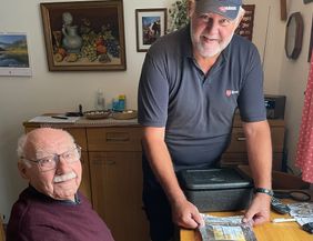 Der Senior sitzt am Tisch, während der Malteser Menüservice-Mitarbeiter die Menüschale für ihn öffnet.