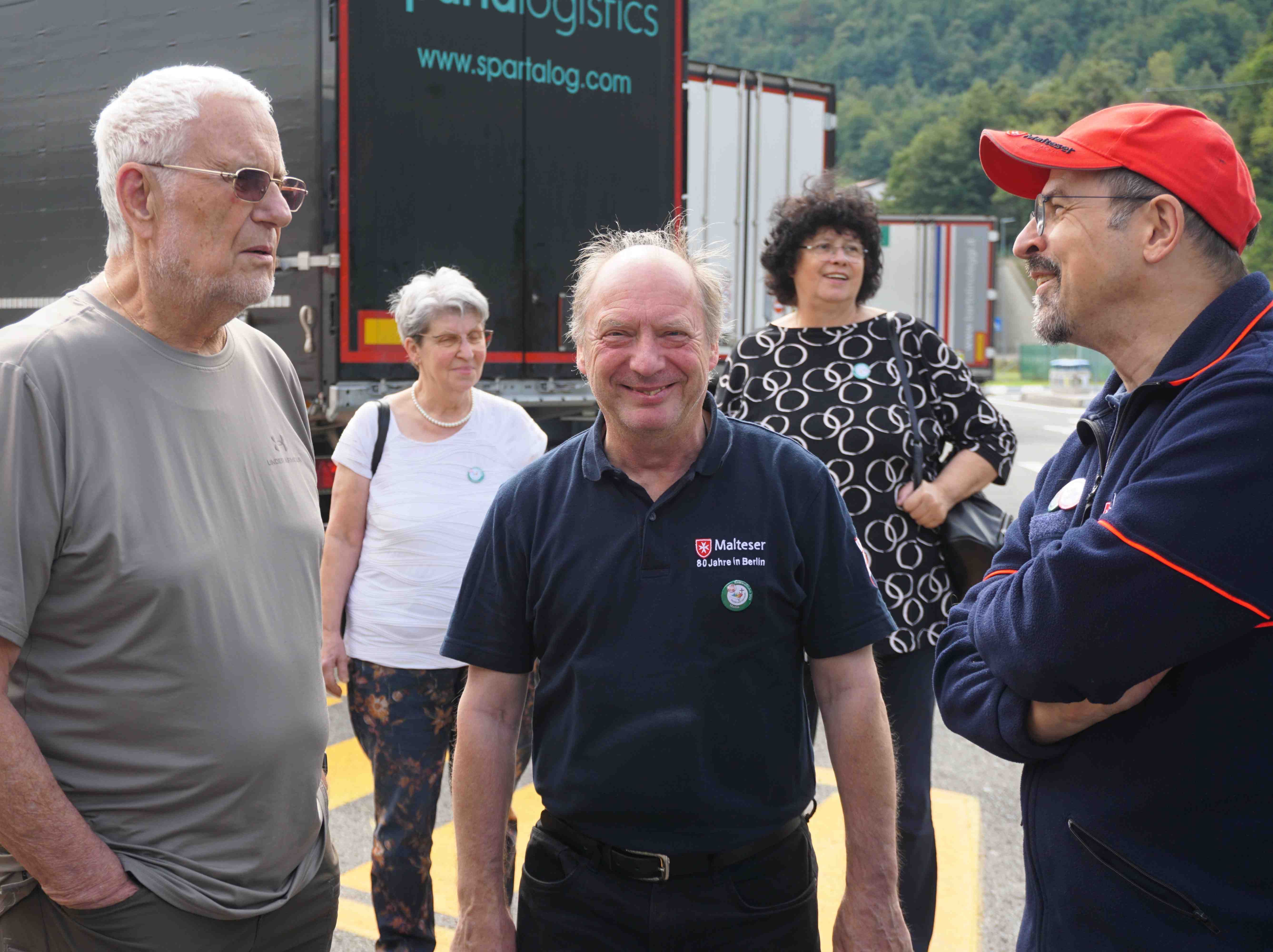 Fünf Personen stehen im Freien auf einem Parkplatz vor Lkw-Aufliegern, eine trägt ein blaues Poloshirt mit Malteser-Logo, eine andere eine rote Kappe und Jacke.