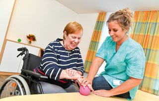 Eine Frau im Rollstuhl bei einer Übung mit einem Ball bekommt Unterstützung von einer Pflegerin
