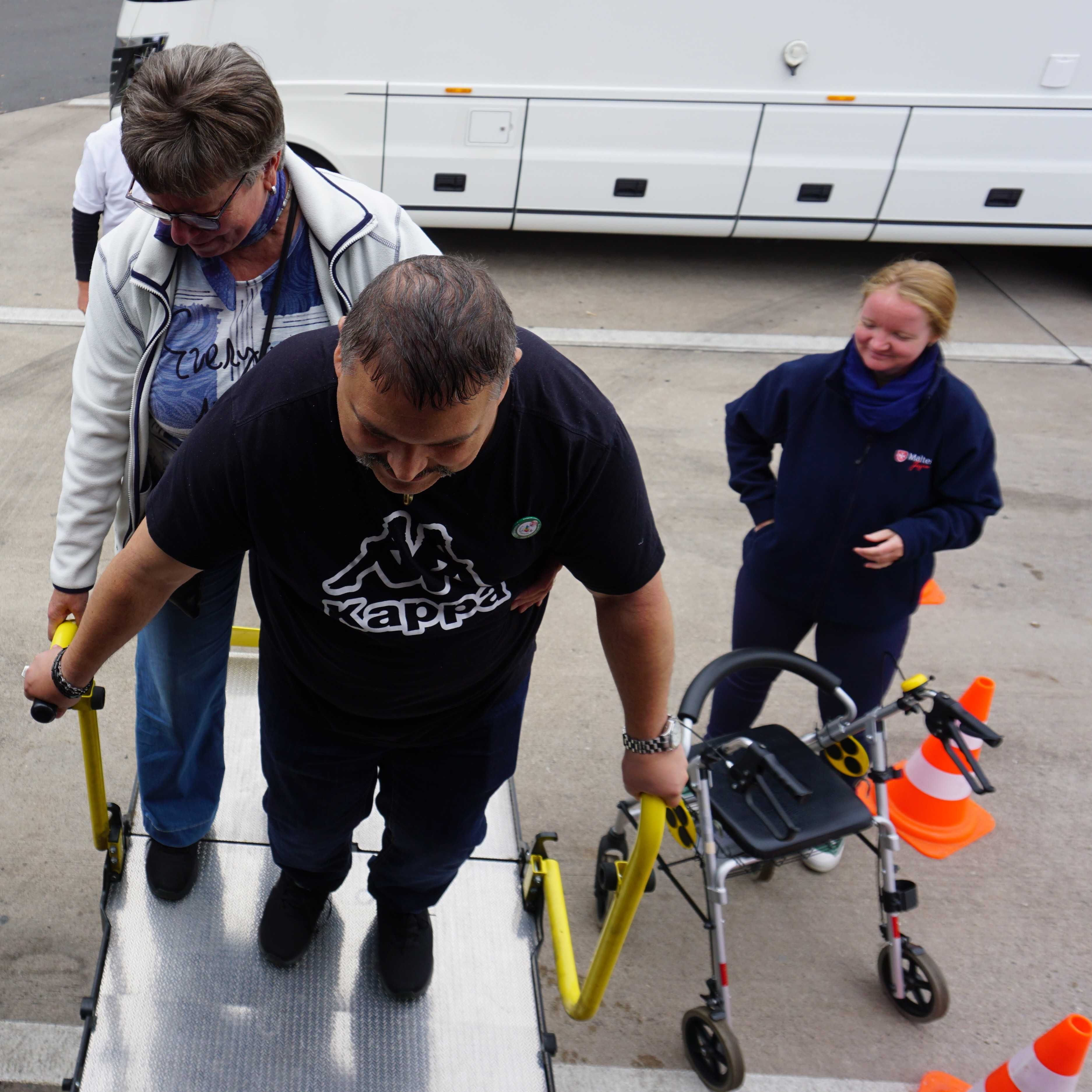 Person steigt mit Hilfe von einem Geländer eine Rampe hinauf, daneben steht ein Rollator und eine weitere Person in dunkler Kleidung.