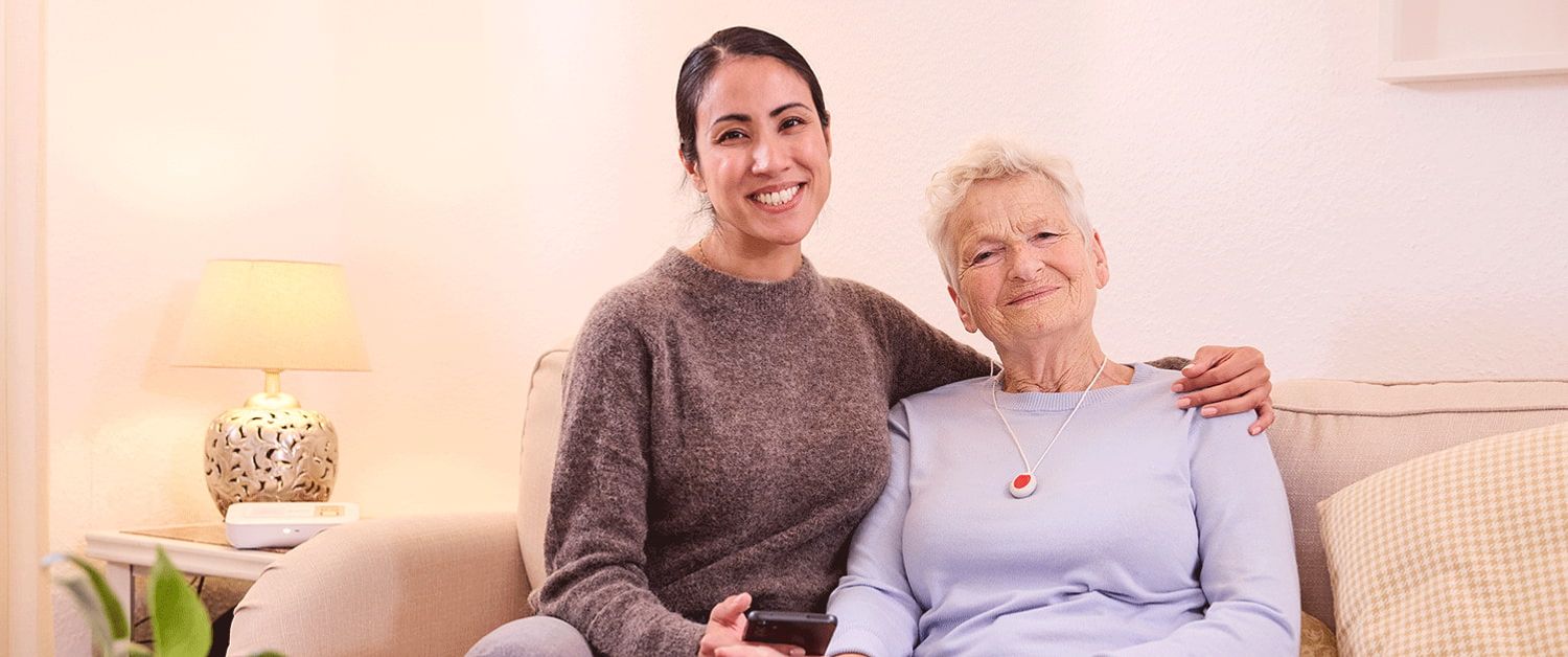 Eine ältere Frau trägt den Hausnotrufknopf als Kette und sitzt mit einer jungen Frau in einem Wohnzimmer.