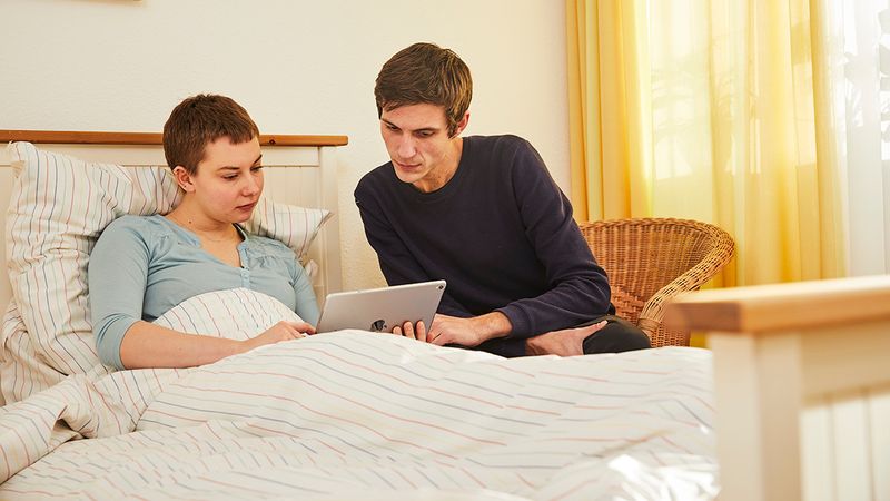 Eine Frau in einem Bett und ein in einem Korbstuhl sitzender Mann schauen auf ein Tablet. 