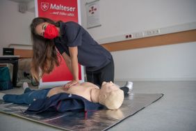 Eine Erste Hilfe Ausbilderin der Malteser macht die Herz-Lungen-Wiederbelebung vor. Aus hygienischen Gründen trägt sie eine Mund-Nasen-Bedeckung. (Foto Malteser/Georg Seeger)