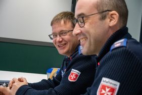 Zwei Männer in Malteser Jacke sitzen nebeneinander und lächeln