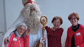 Eine große Person in traditionellem Nikolausgewand steht in der Mitte. Das Gewand ist weiß, dazu trägt die Person eine rote Mitra mit goldenen Verzierungen und hält einen goldfarbenen Bischofsstab in der rechten Hand. Über dem linken Arm hängt ein brauner Jutesack. Links und rechts vom Nikolaus stehen drei Senioren des Malteser Besuchsdienstes in festlicher Kleidung, die sich eng an ihn stellen. Die Gruppe befindet sich in einem Raum mit hellem Holzboden und weißer Wand im Hintergrund.