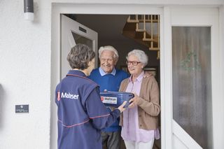 Älteres Ehepaar steht an der Haustür und bekommt von einem Malteser eine Box überreicht