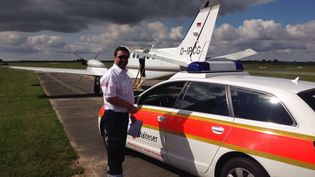 Malteser Mitarbeiter steht neben einem Malteser Wagen vor einem Flugzeug.