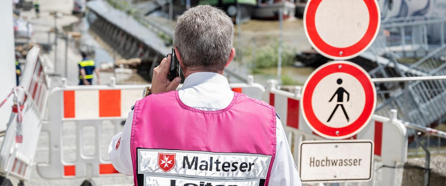 Ein Mann mit einer Weste mit Malteser-Logo telefoniert vor einer abgesperrten Straße