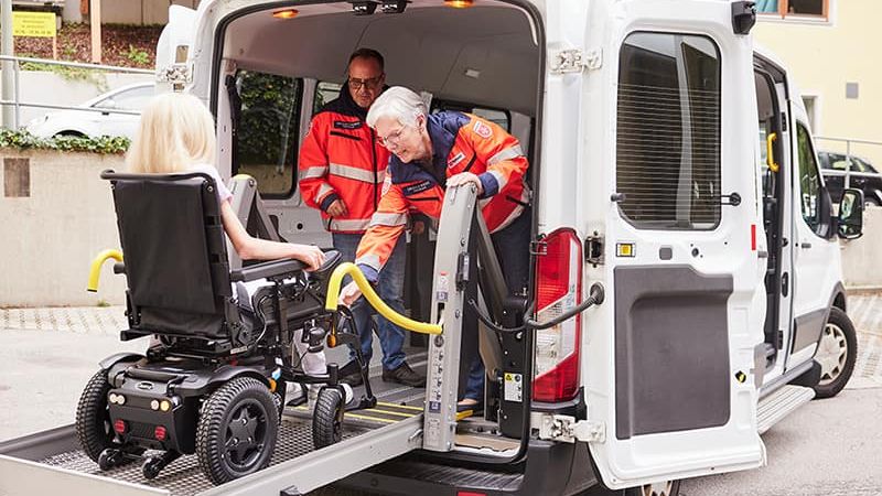 Zwei Mitarbeiter der Malteser helfen Frau im Rollstuhl in den Wagen