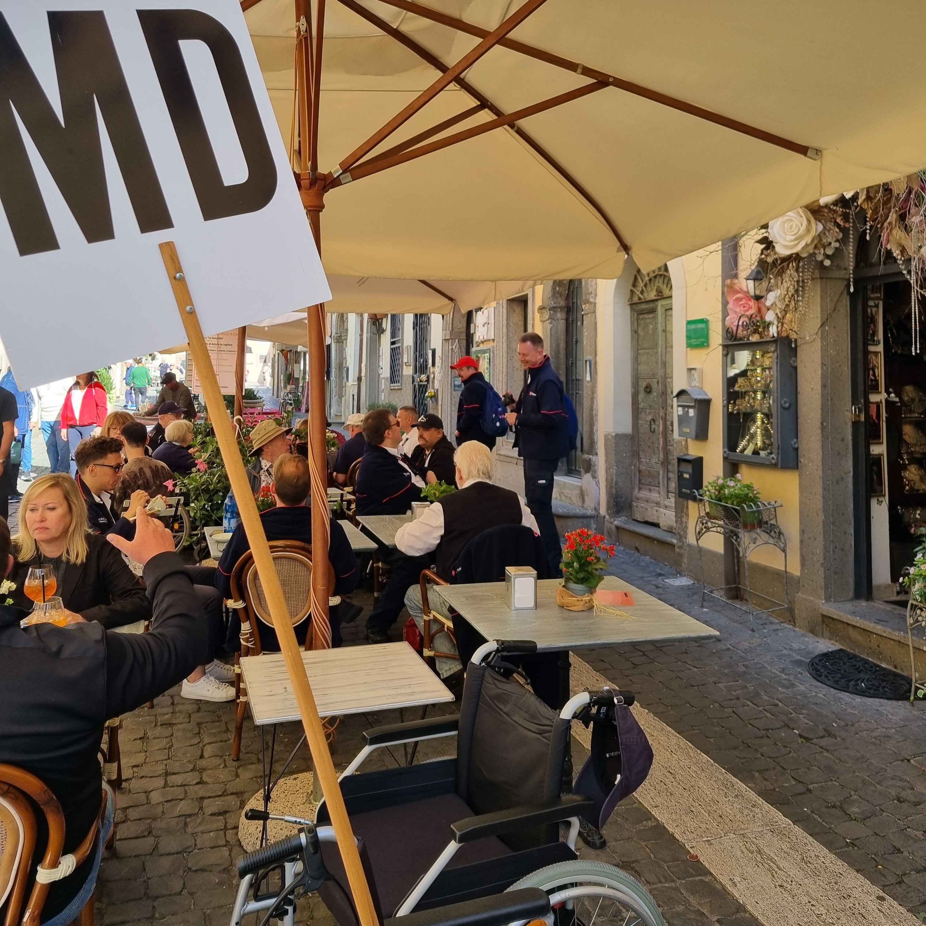 Straßencafé mit Tischen, Stühlen und Sonnenschirmen, eine Person hält ein Schild mit der Aufschrift 'MD'.
