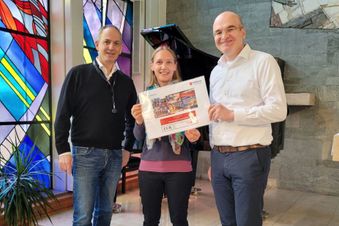 Drei Personen in einer Kapelle mit bunten Fenstern halten einen Spendenscheck.