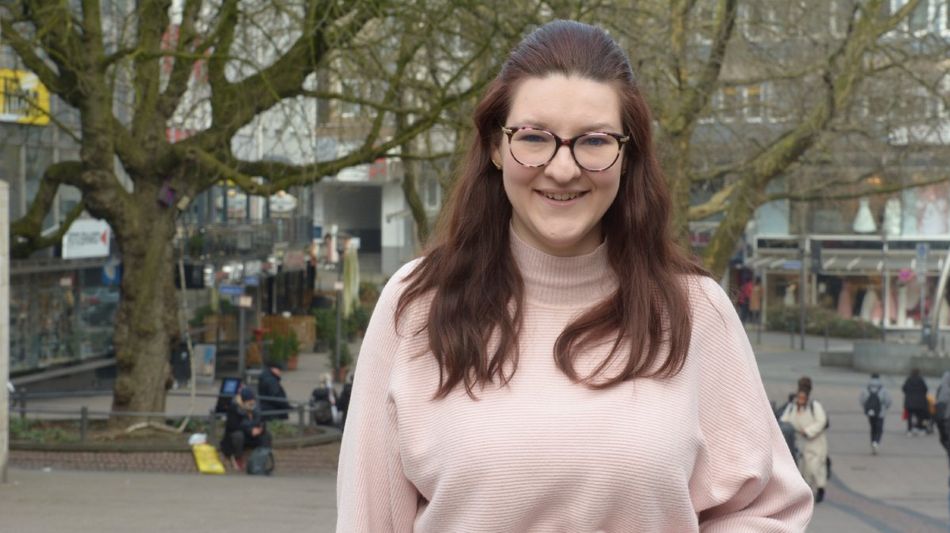 Eine lächelnde junge Frau mit Brille in einer Fußgängerzone.