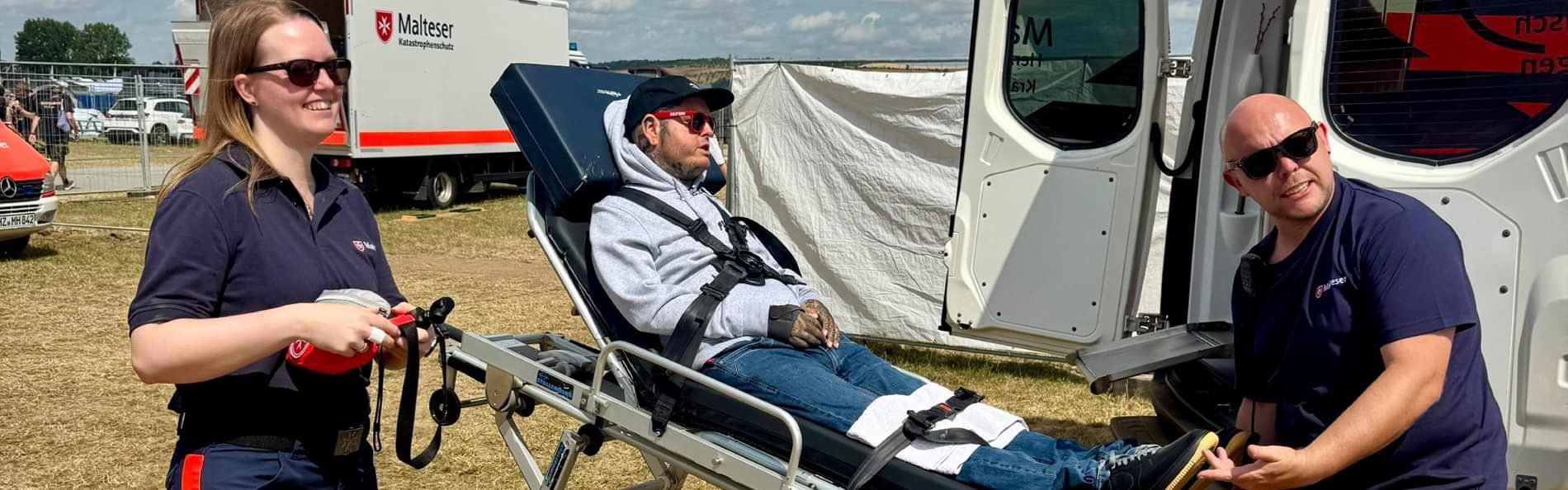 Ein Mann mit Baseballmütze und Sonnenbrille auf einer Rollbahre und zwei junge Menschen in Einsatzkleidung der Malteser auf einem Festivalgelände. 