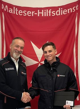 Zwei Männer stehen lächelnd vor einer Malteser-Fahne mit Handschlag. Foto: Malteser