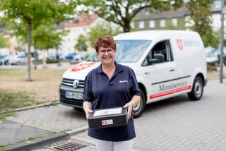 Mitarbeiterin des Malteser Menueservice trägt eine Warmhaltebox und lächelt in die Kamera.