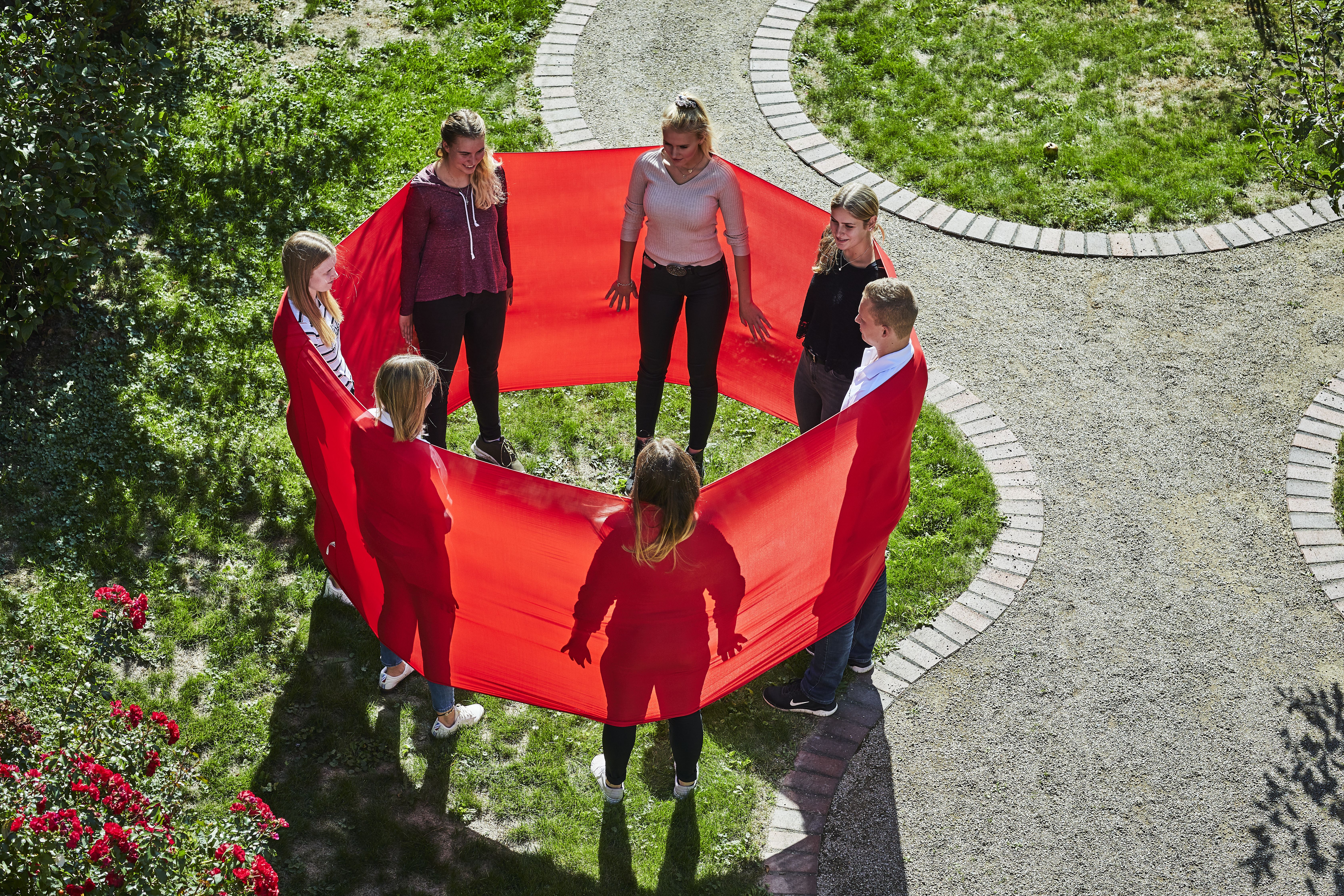 Sieben junge Menschen stehen im Freien in einem Kreis und haben ein rotes Band um sich gespannt.