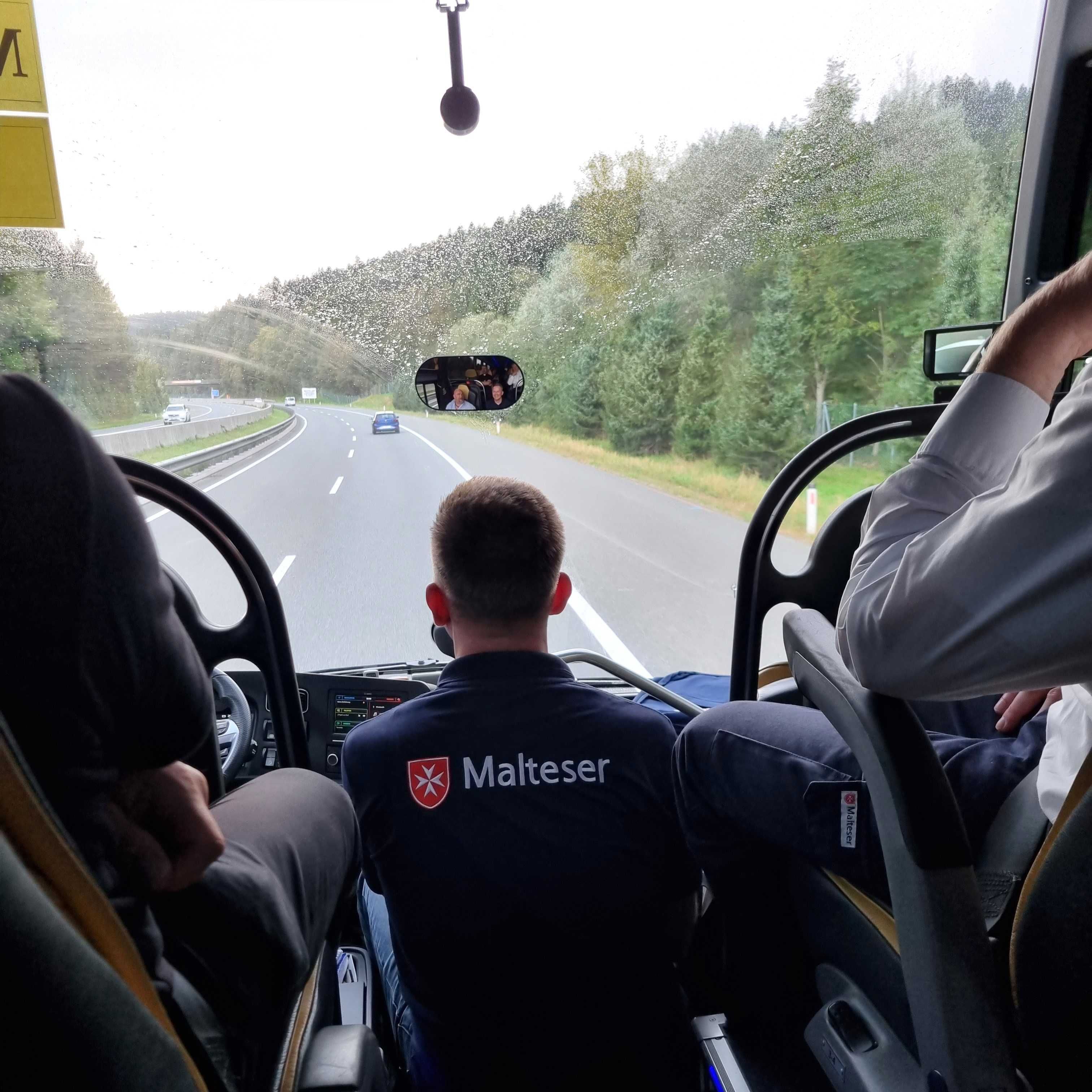 Rückenansicht eines Mannes mit Malteser-Logo auf dem Shirt, sitzend im Bus mit Blick auf die Straße und Fahrer.