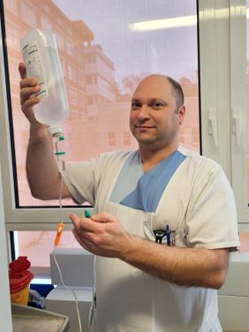 Pflegekraft in weißer Kleidung hält Infusionsflasche hoch und prüft den Schlauch vor einem Fenster mit Tageslicht.