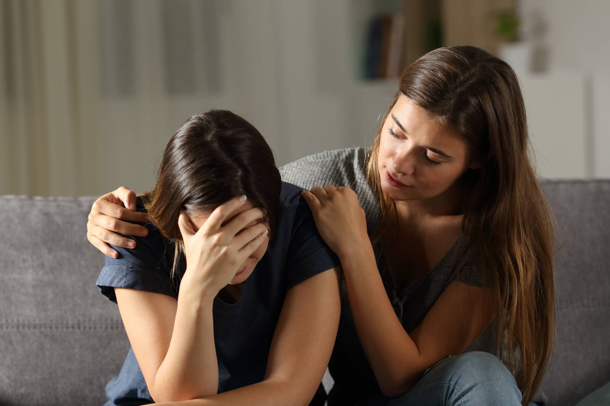 Eine junge Frau umarmt und tröstet einen andere junge Frau sitzend auf einem Sofa