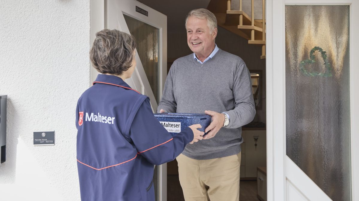 Eine Mitarbeiterin des Malteser Menüservice übergibt eine Mahlzeit an der Tür. 