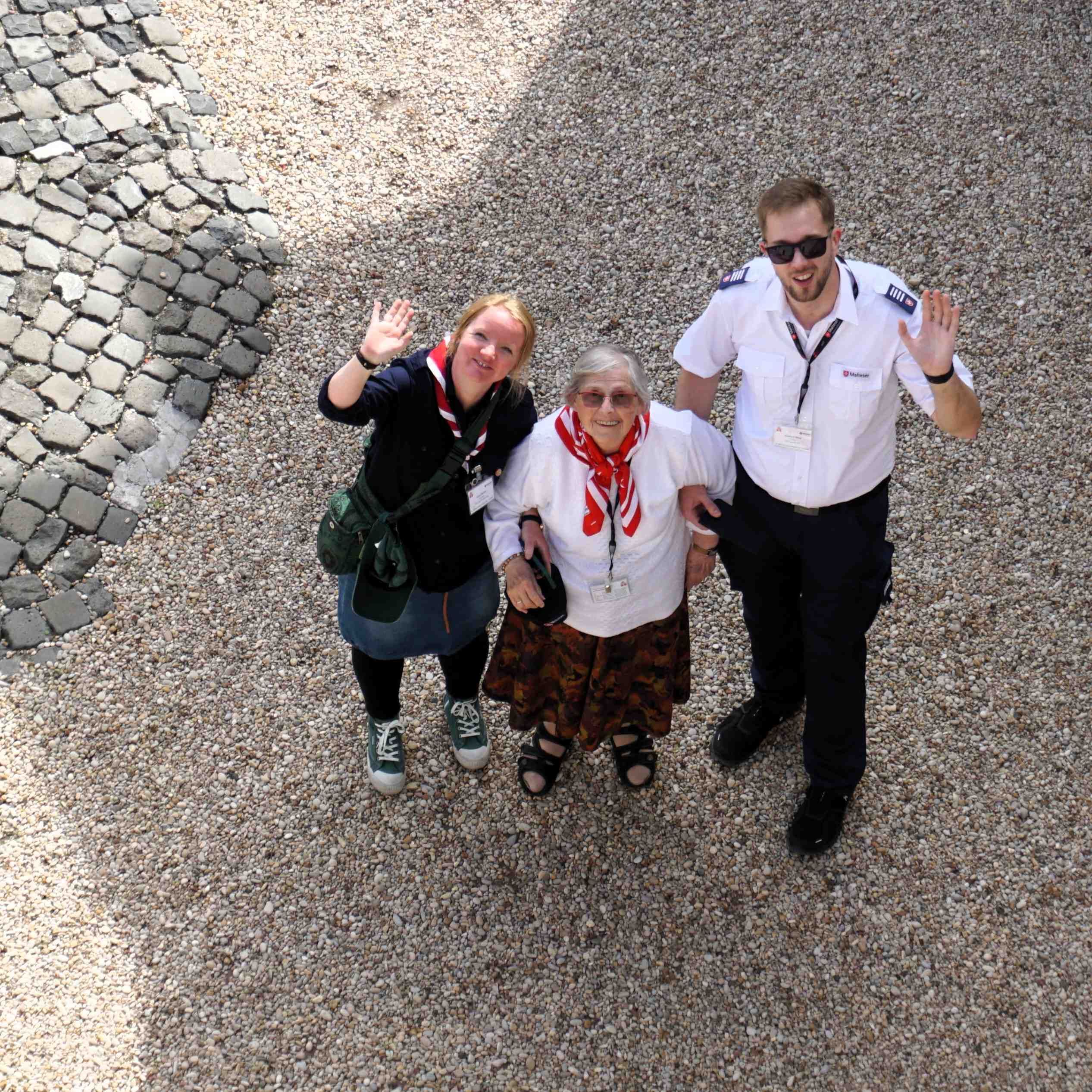 Drei Personen stehen auf gepflastertem Boden, zwei winken, eine trägt ein weißes Hemd mit rotem Halstuch.