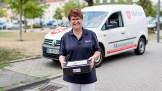 Mitarbeiterin des Malteser Menueservice trägt eine Warmhaltebox und lächelt in die Kamera.