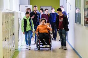 In einem Schulgang ist eine Gruppe von Schülerinnen und Schülern auf dem Weg ins Klassenzimmer. Inmitten der Gruppe ein junger Mann im Rollstuhl, der von einem Malteser Helfer begleitet wird. und in Begleitung eines Malteser helfers.