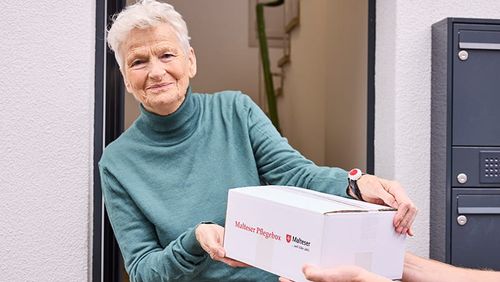 Eine ältere Frau mit kurzen grauen Haaren nimmt an der Haustür ein Päckchen mit der Malteser Pflegebox entgegen.