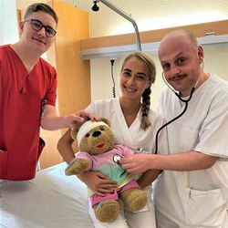 Drei Pflegekräfte versorgen auf einem Klinikbett spielerisch einen Teddybären mit Verband und Stethoskop