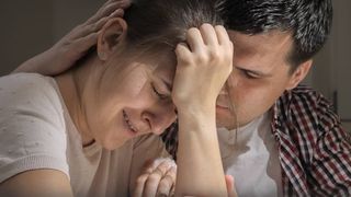 Ein Mann tröstet eine weinende Frau, die am Tisch sitzt. 