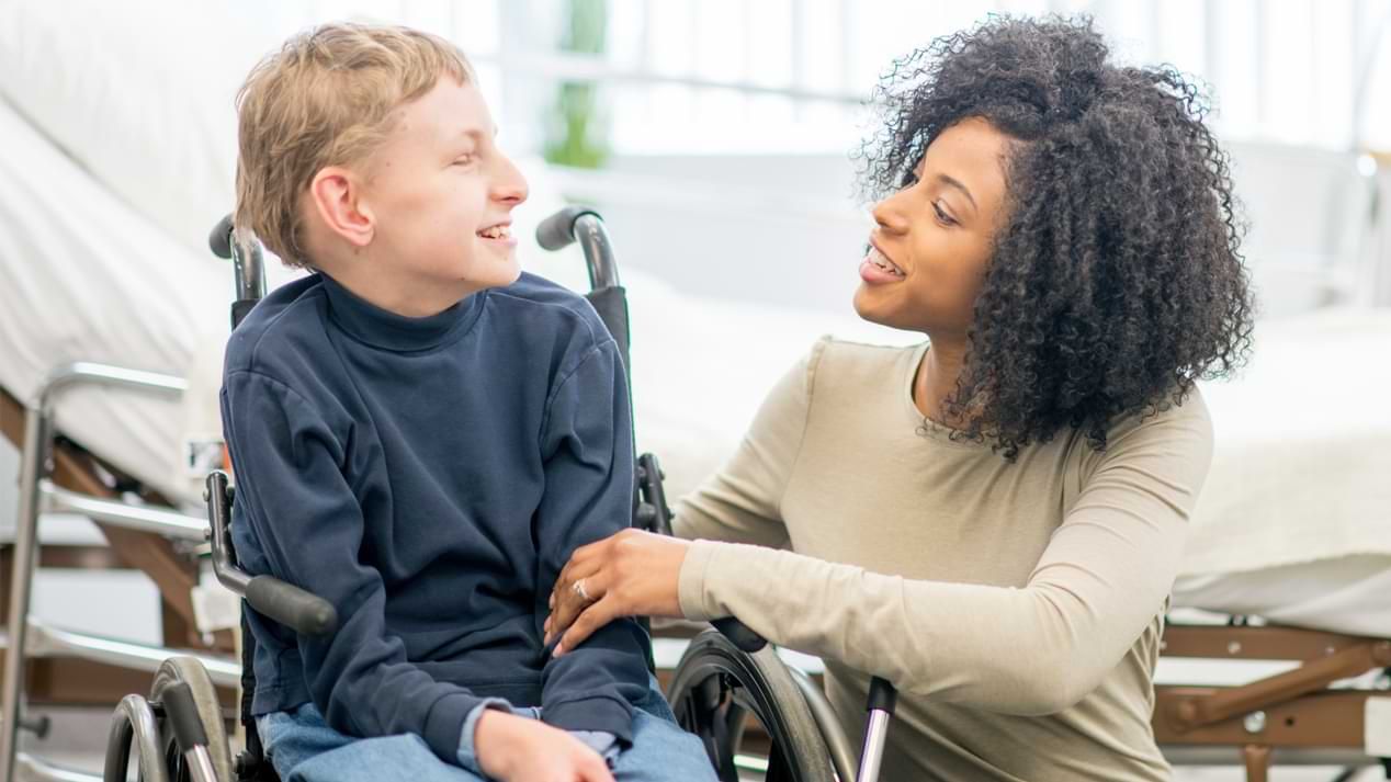 Ein Junge im Rollstuhl und eine knieende junge Frau in einem Krankenzimmer.