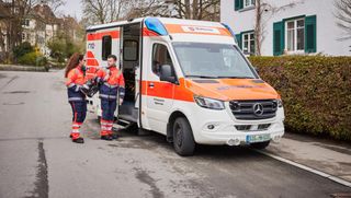 Rettungshelfer und Rettungshelferin der Malteser vor einem Rettungswagen aus der Ferne