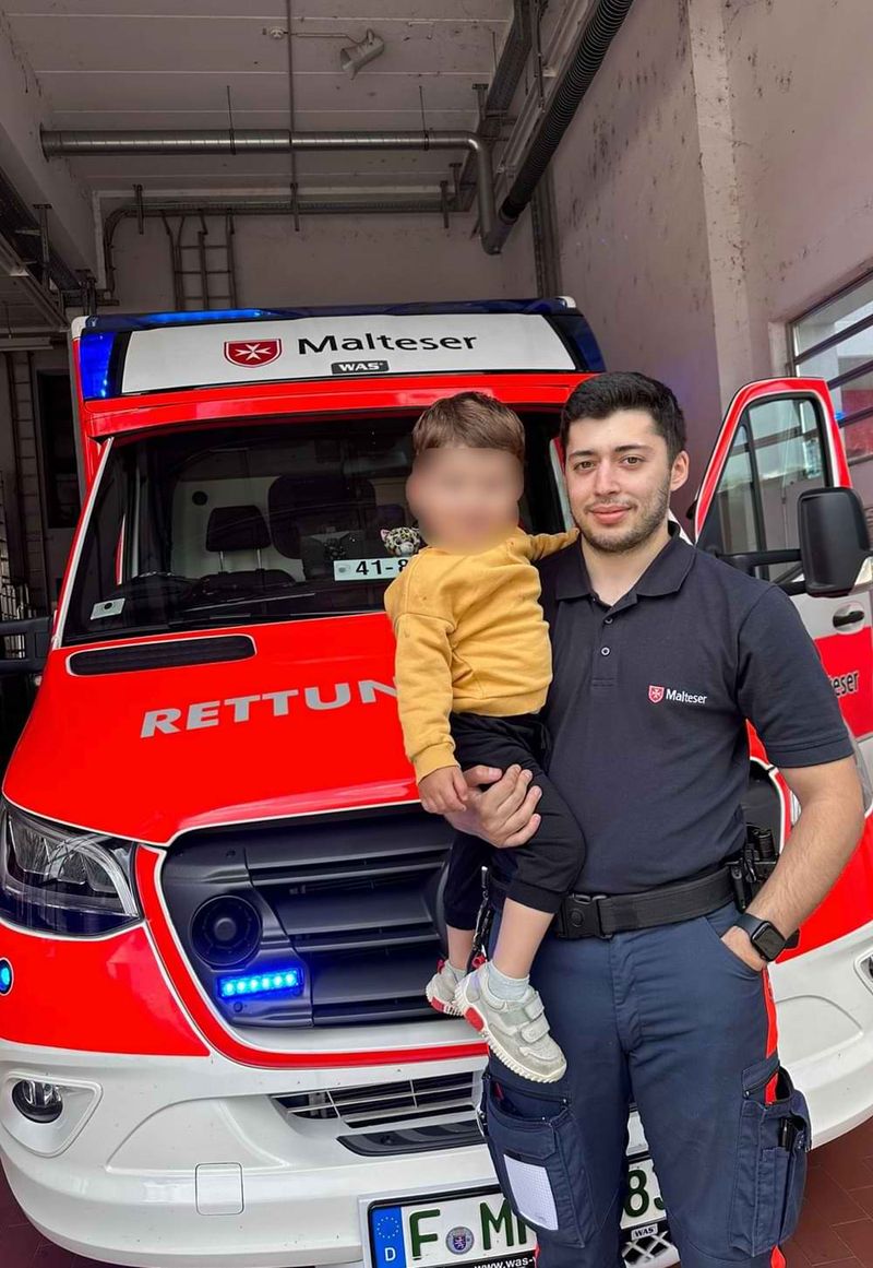 Ein Rettungssanitäter der Malteser vor einem RTW mit einem kleinen Jungen auf dem Arm.