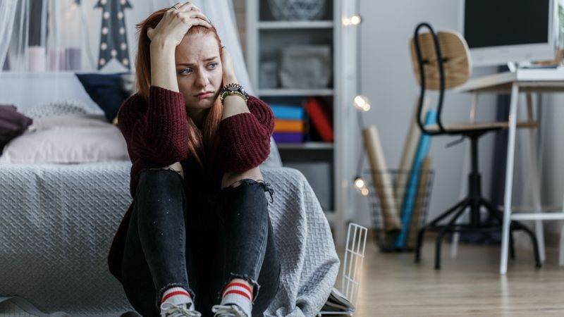 Junge Frau hockt deprimiert auf dem Fußboden eines Schlafzimmers.