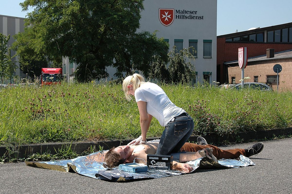 Auf dem Bild ist eine junge Frau zu sehen, die bei einer Person auf dem Boden liegend Herzdruckmassage macht.